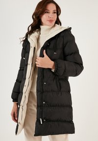 Puffer coat with a black exterior and cream interior. Features a hood, zippered pockets, and elastic cuffs. Textured fabric with quilted design.