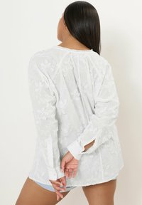 Blusa bianca ricamata con maniche lunghe, caratterizzata da motivi floreali, polsini con bottoni e un tessuto trasparente testurizzato. Vista dal retro.