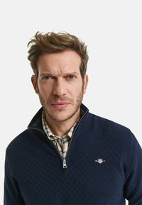 Marineblauer Reißverschluss-Pullover mit strukturiertem Muster, darunter ein kariertes Hemd und ein Logo auf der Brust. Silberne Reißverschluss-Details.