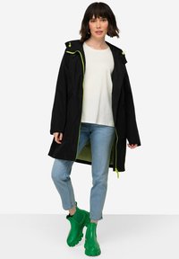 Longue veste noire avec un intérieur vert fluo et des fermetures éclair, dotée d'une capuche et de poches latérales. Portée avec un jean bleu clair et des bottes en caoutchouc vertes.