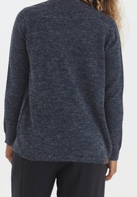 Žena oblečená v tmavě modrém chlupatém svetr a černých kalhotách, zobrazená zezadu s částečně viditelnými kudrnatými vlasy.