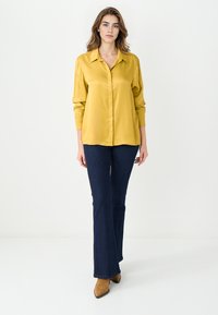 Koszula z długim rękawem, musztardowo-żółta, zapinana na guziki, z kołnierzykiem, w połączeniu z ciemnoniebieskimi dżinsami w fasonie dzwonów i jasnobrązowymi, czubkami w kształcie szpica, botkami za kostkę.