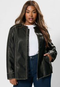 Giubbotto di pelle nera con zip frontale, colletto rialzato e cuciture visibili. Indossato sopra una t-shirt bianca e jeans blu a vita alta.