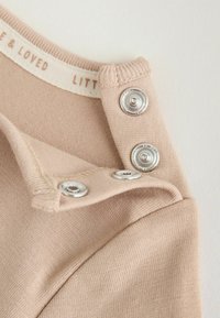 Gros plan sur un tissu beige avec des boutons-pression argentés et un texte partiel sur l’étiquette intérieure indiquant "LOVE & LOVED LITT".