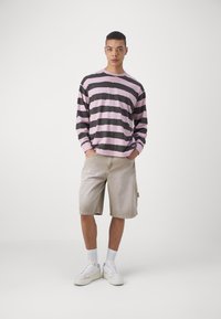Långärmad skjorta med rosa och svarta ränder, kombinerad med beige shorts. Modellen har på sig vita sneakers och synliga vita strumpor.