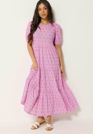 Femme aux longs cheveux foncés portant une robe maxi rose à volants avec des pois noirs et des sandales blanches à enfiler sur un fond uni.