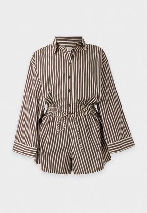 Mujer con camisa de botones de rayas verticales marrón y blanca y pantalones cortos a juego, con calcetines blancos y zapatos marrón oscuro, de pie frente a un fondo blanco.