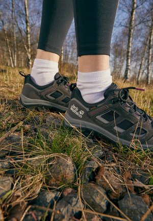 Person trägt schwarze Jack Wolfskin Wanderschuhe und weiße Socken und steht auf felsigem Gras in einem bewaldeten Gebiet mit Birken.