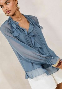 Light blue, sheer blouse with ruffled neckline and sleeves, featuring a layered design and subtle texture. Pairs well with white shorts.