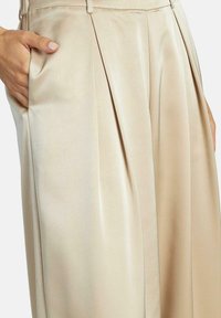 Pantalons plissés beiges en satin, dotés de poches latérales et d'une texture lisse, avec une taille ajustée pour un look cintré.