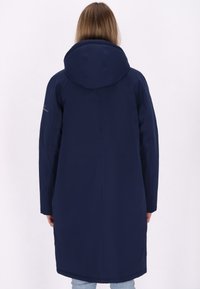 Persoon met lang haar in een marineblauwe hooded coat, staand met de rug naar de camera tegen een effen lichte achtergrond.