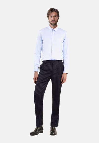 Hellblaues Langarmhemd mit spitzem Kragen, kombiniert mit dunkel gestreiften Hosen und schwarzen Knöchelschuhen. Schlichtes Design, strukturierte Passform.