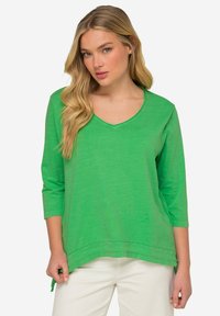 Grünes Langarmoberteil mit V-Ausschnitt, aus leichtem Stoff gefertigt. Es hat eine lockere Passform und einen abgerundeten Saum mit seitlichen Schlitzen.
