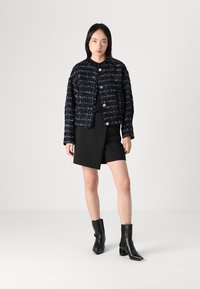 Navy tweed jasje met een gestructureerd geweven patroon, ronde kraag en knoopsluiting, gecombineerd met een zwarte rok en puntige enkellaarzen.