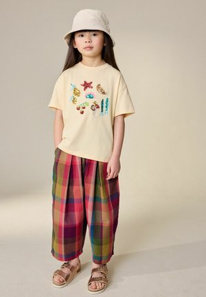 Young girl wearing a beige sequin-embellished t-shirt, colorful plaid wide-leg pants, beige bucket hat, and metallic buckle sandals standing.