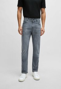 Jeans in denim grigio con taglio dritto, tasche anteriori e chiusura con bottone, abbinati a sneaker bianche e una maglietta nera a maniche corte.