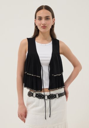 Femme portant un top blanc sans manches, un gilet noir superposé avec des clous argentés, une jupe blanche, une ceinture noire cloutée et de grandes boucles d'oreilles.