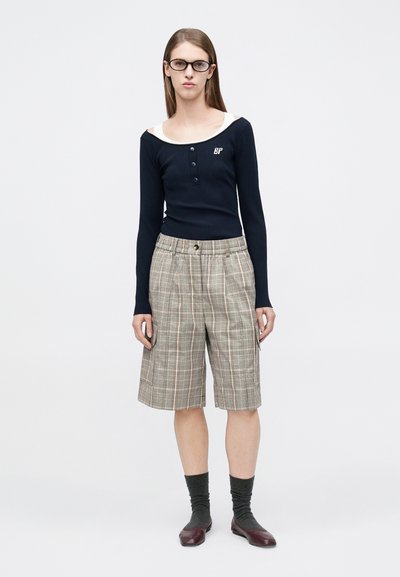 Μπλε ναυτικό μακρυμάνικο τοπ με ριμπ ύφανση, κρεμ γιακά και κουμπιά, συνδυασμένο με μπεζ καρό σορτς με πορτοκαλί και πράσινες ρίγες και καφέ παπούτσια.