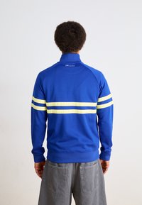 Mėlynas sportinis džemperis su aukštu apykakle, papuoštas geltonomis horizontaliomis juostelėmis. Pagamintas iš lygaus audinio, turi prigludusį dizainą ir raukinėtas rankogalius.