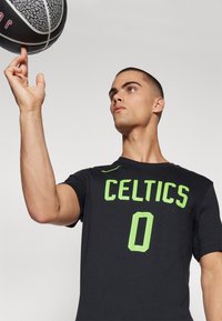 Must have musternõudega must sportlik T-särk, millel on roheline „CELTICS“ kiri ja number „0“. Tekstuuriga kangas ja keha jäljendav lõige. Mängija tasakaalustab korvpalli.