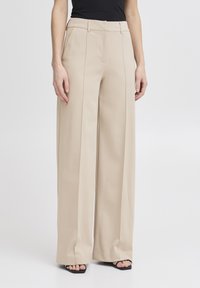 Beige kalhoty s širokými nohavicemi a hladkou texturou, se sklady na přední straně, poutky na opasek a přiléhavým střihem, v kombinaci s černými sandály.