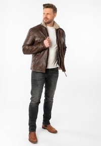 Braune Lederjacke mit Schaffellkragen, mit Fronttaschen und Reißverschluss, getragen über einem hellen Hemd, kombiniert mit dunklen Jeans und braunen Stiefeln.