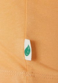 Oranžinė medžiaga su siauru reljefu ir balta etikete, ant kurios pavaizduotas žalias lapo logotipas. Rodyti tvarkingą siuvimą aplink kraštus.