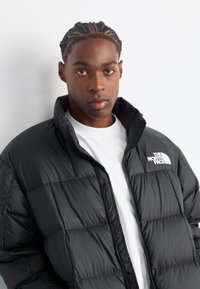 Crna puffer jakna s kvadratnim dezenom i višim ovratnikom. Logo The North Face u bijeloj boji prikazan je s lijeve strane. Bijela majica ispod.