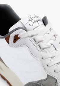 Zapatilla blanca con acentos de ante gris y marrón, tejido texturizado, cierre de cordones y logo característico en la lengüeta, con suela lisa.