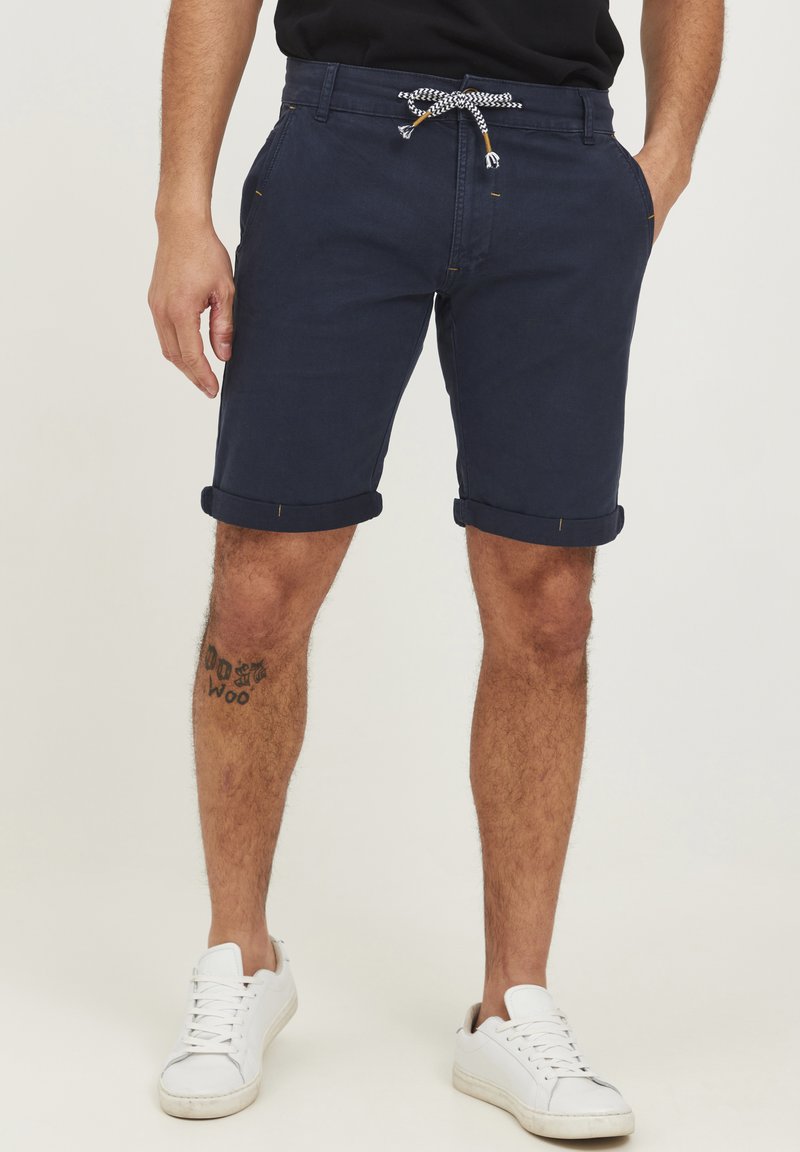 Marineblå bomullscargo shorts med oppbrettede ermer, snor i livet og lommer. Har gule sømdetaljer. Kombinert med hvite joggesko.