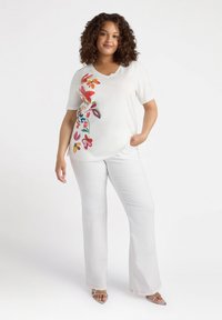Top blanco de manga corta con bordado floral colorido en el lado izquierdo, combinado con pantalones blancos acampanados y sandalias de tacón plateadas.