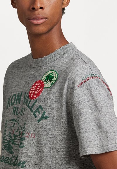 Harmaa puuvillainen t-paita, jossa on vihreitä ja punaisia graafisia painatuksia, pyöreät tikkaukset ja koristeompeleet olkapäässä.