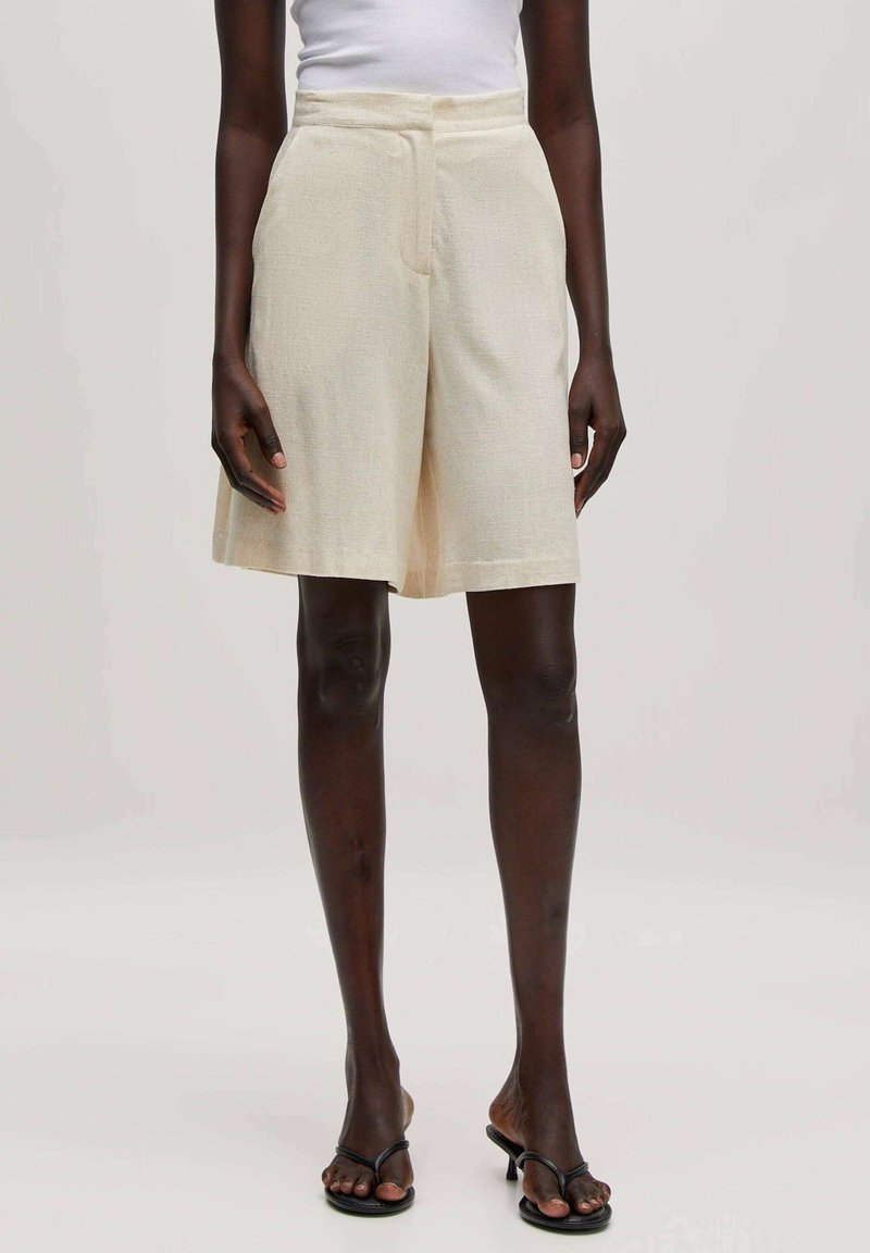 Beige linnen shorts met een hoge tailleband, voorplooien en zijzakken; de zoom valt boven de knie met een lichte, textuurafwerking.