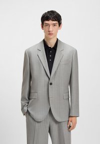 Young man wearing light gray pinstripe suit with black polo shirt, standing with one hand in pocket against white background.