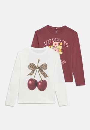 Deux chemises à manches longues, une blanche avec des cerises et un nœud léopard, une bordeaux avec des fleurs et le texte "MOMENTS", à plat sur fond blanc.