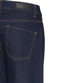 Jean en denim bleu foncé avec coutures orange, comprenant un patch arrière avec le nom de la marque, et deux poches arrière avec des coutures renforcées.