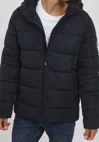 Marineblaue Steppjacke mit Reißverschluss vorne, Kapuze und gesteppten Einsätzen. Verfügt über seitliche Reißverschlusstaschen und ein Logo am linken Ärmel.