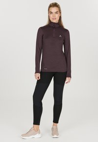 Bordeaux langærmet top med kvart lynlås, tekstureret stof og et lille logo. Sammen med sorte tætsiddende leggings og lyse atletik sko.