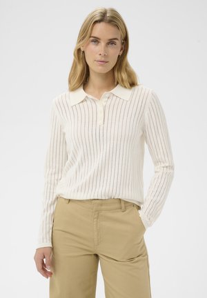 Junge Frau mit blonden Haaren trägt ein weißes langärmliges Strick-Polo-Shirt mit vertikalen Streifen und beigen Hosen und posiert mit einer Hand in der Tasche.