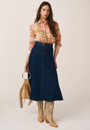 Mujer con una camisa de botones estampada metida dentro de una falda midi de mezclilla oscura con cinturón, sosteniendo un bolso marrón con flecos y usando botas beige con estampado floral.