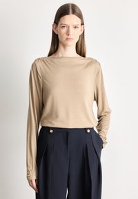 Beżowy top z długim rękawem i dekoltem typu łódka, wykonany z miękkiego materiału, z marszczonymi detalami na ramionach. W zestawie granatowe spodnie z ozdobnymi złotymi guzikami.