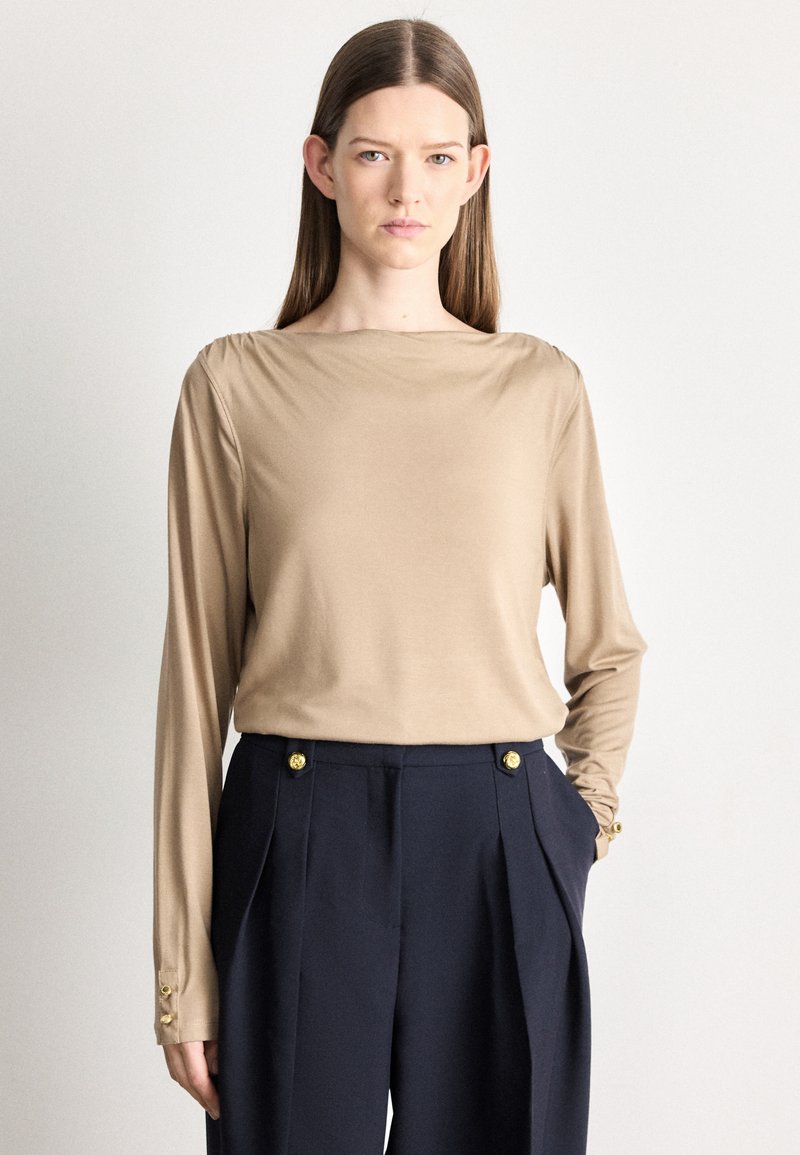 Beżowy top z długim rękawem i dekoltem typu łódka, wykonany z miękkiego materiału, z marszczonymi detalami na ramionach. W zestawie granatowe spodnie z ozdobnymi złotymi guzikami.