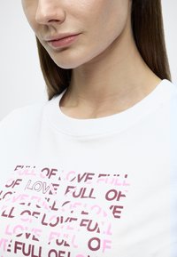 Λευκή φούτερ μπλούζα με το κείμενο «FULL OF LOVE» σε ροζ και βουργουνδί χρώματα, με λαιμόκοψη crew και κοντά μανίκια. Το κείμενο έχει διαφορετική διαφάνεια.