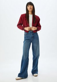 Burgundy cropped jakke med dobbelte knapper, parret med højtaljede, vidde blå jeans og hvide sneakers med brune såler.