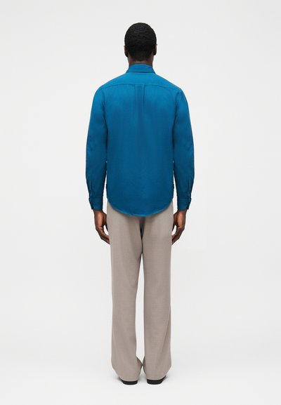 Τυρκουάζ μπλούζα με μακριά μανίκια και καμπύλη λαιμόκοψη, συνδυασμένη με ανοιχτό γκρι παντελόνα με πλατιά μπατζάκια. Η μπλούζα έχει μανσέτες με κουμπιά και άνετη εφαρμογή.