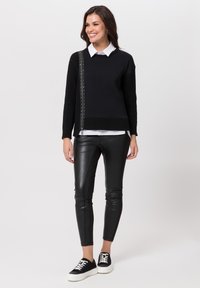 Schwarzer Pullover mit strukturierten Ärmeln und silbernen Nieten, kombiniert mit glänzenden schwarzen Lederleggings und schwarzen Sneakern mit weißen Sohlen.