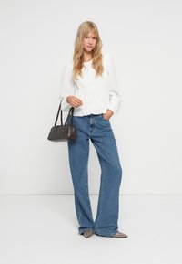 Hvid, tekstureret bluse med lange puffærmer, parret med højtaljede, blå brede jeans. Modellen holder en sort, rektangulær håndtaske.