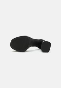 Chaussure à talon haut noire avec une tige en cuir lisse et un talon carré. La semelle en caoutchouc texturé présente des rainures horizontales pour une meilleure adhérence.