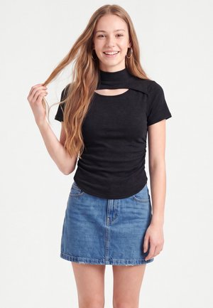 Jeune femme aux longs cheveux souriante, portant des créoles, un haut noir à manches courtes avec une découpe et une jupe en jean bleue, tenant ses cheveux d'une main.