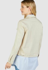 Helle beige Jeansjacke mit langen Ärmeln, slim fit, geknöpften Manschetten und Rückenpasse, getragen über einem blau gestreiften Hemd.
